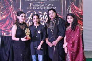 The image captures a proud and celebratory moment at Bangalore Fashion Trainings’ 1st Annual Day & Certification Ceremony. The elegant stage backdrop features the “Restyle Revolution” theme with dramatic red-and-black fashion visuals, creating a glamorous fashion-show atmosphere. The coordinated outfits, trophy, smiles, and stage lighting beautifully reflect achievement, creativity, confidence, and celebration.