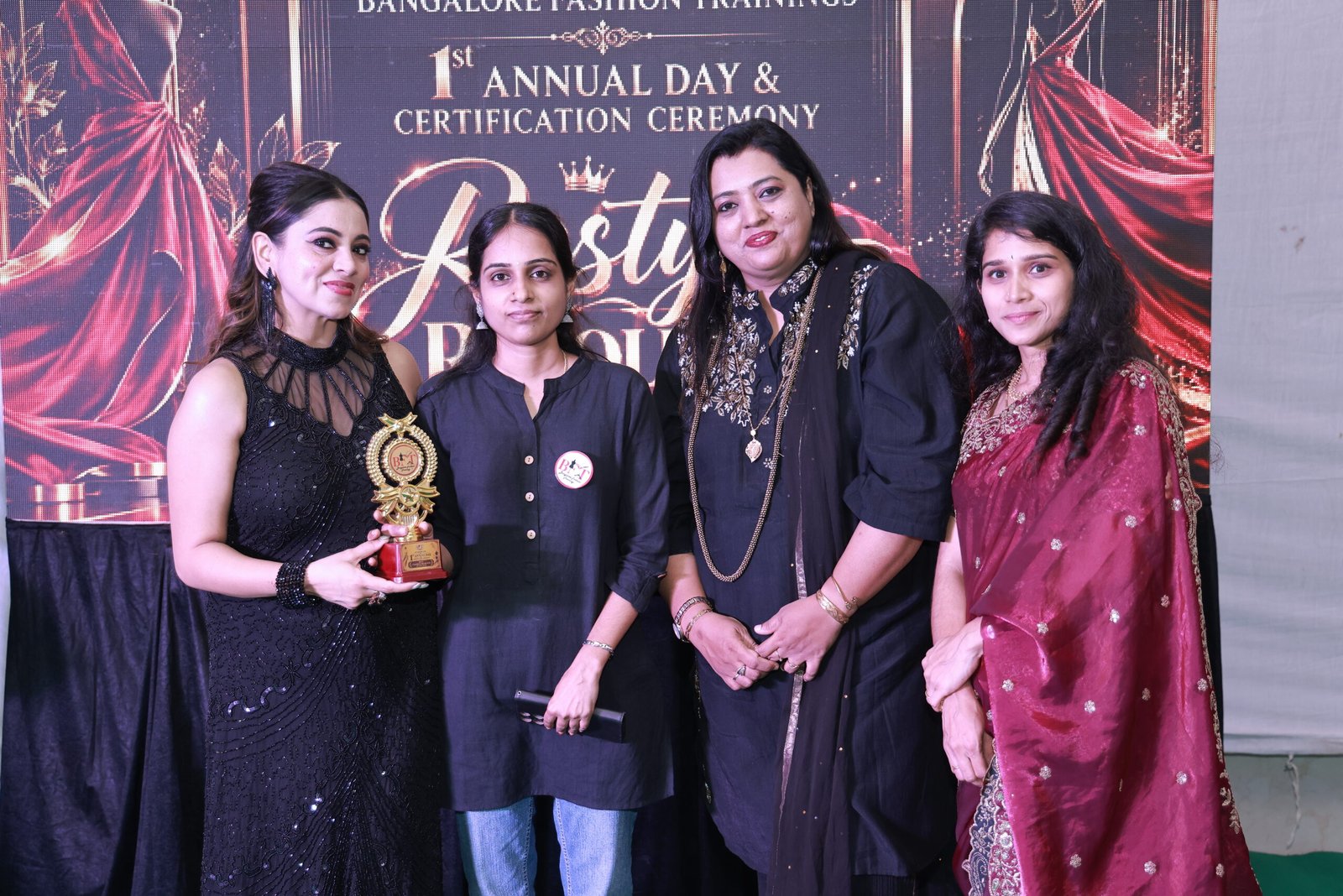 The image captures a proud and celebratory moment at Bangalore Fashion Trainings’ 1st Annual Day & Certification Ceremony. The elegant stage backdrop features the “Restyle Revolution” theme with dramatic red-and-black fashion visuals, creating a glamorous fashion-show atmosphere. The coordinated outfits, trophy, smiles, and stage lighting beautifully reflect achievement, creativity, confidence, and celebration.